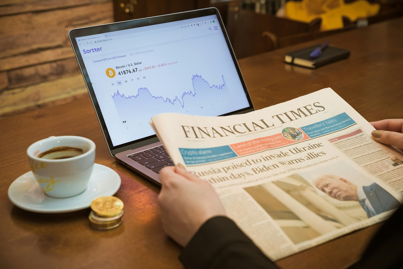 A person reading a Financial Times newspaper beside a laptop showing a Bitcoin price chart, with coffee and a gold coin on the table