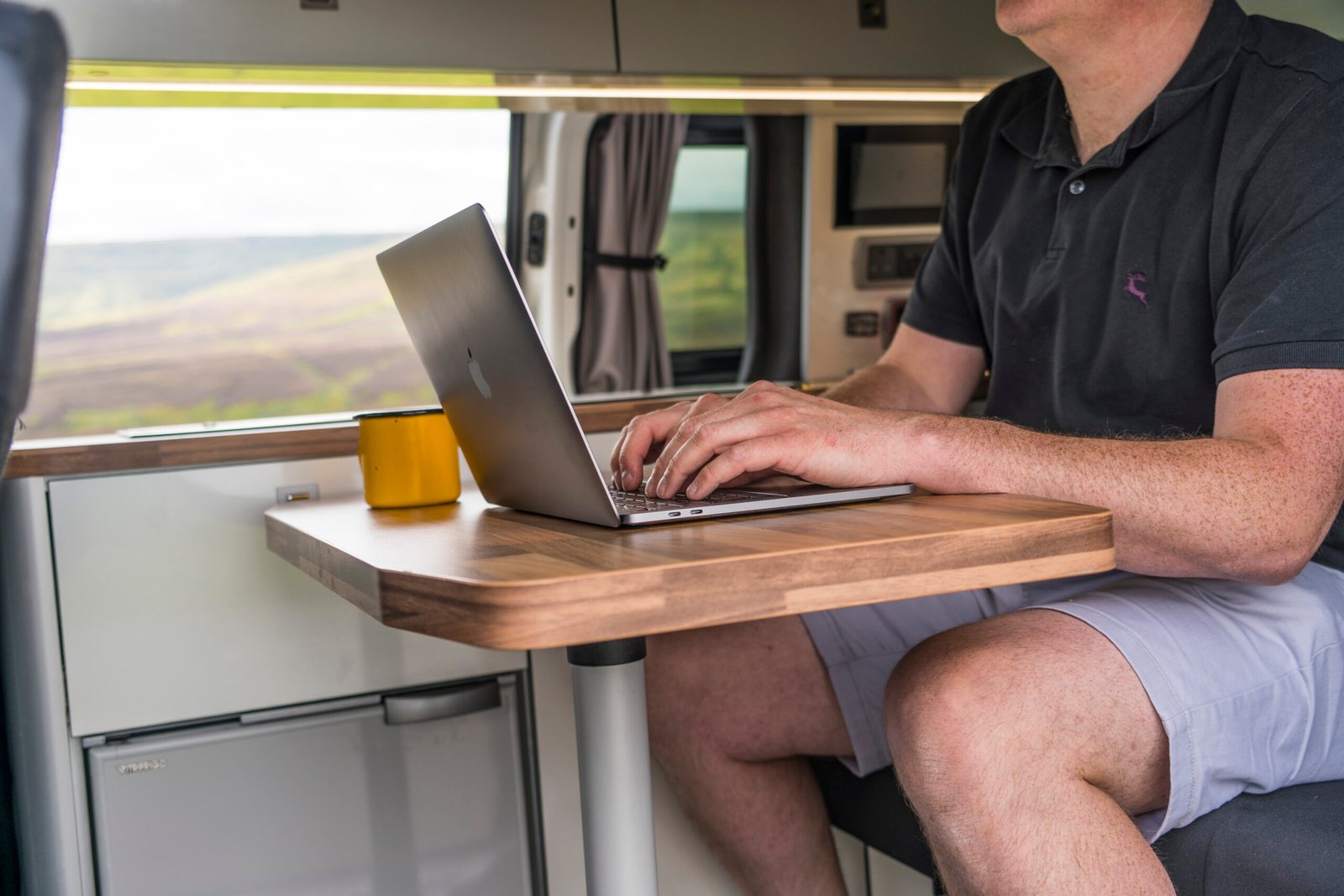 Man typing on a laptop at a fold-out table inside a camper van with a scenic view outside