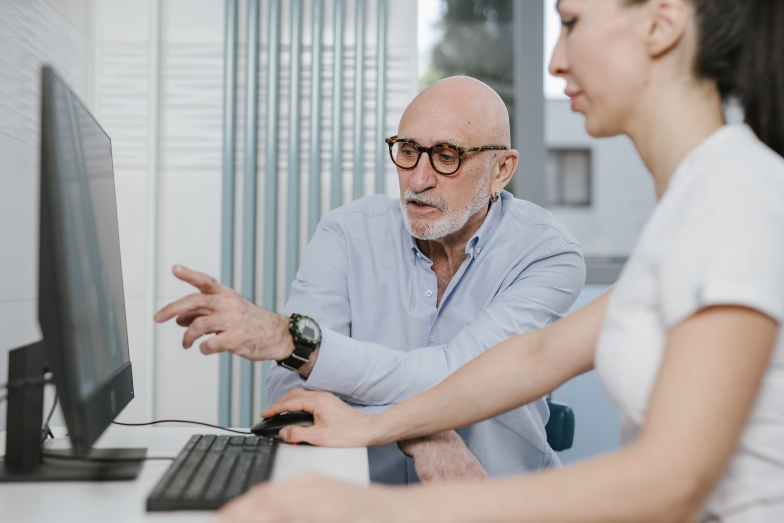 An older man pointing at a computer screen while a younger woman uses the mouse