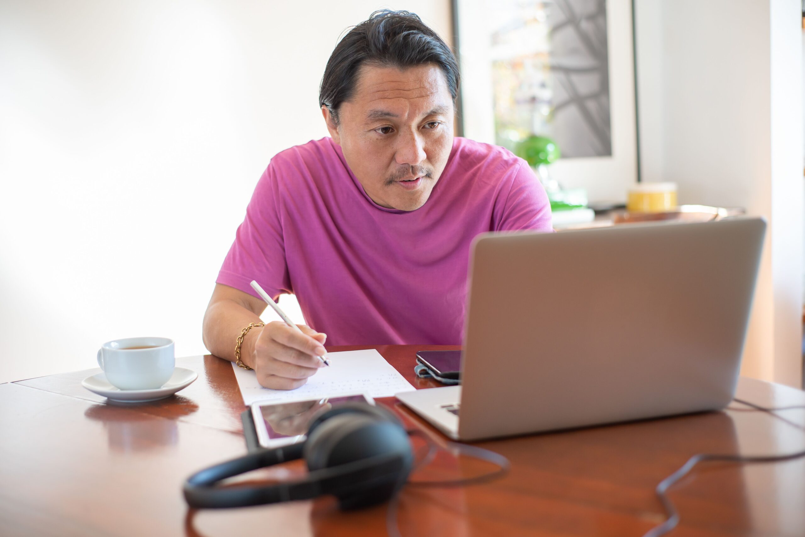 A man writing notes at a desk with a laptop, coffee, and headphones nearby