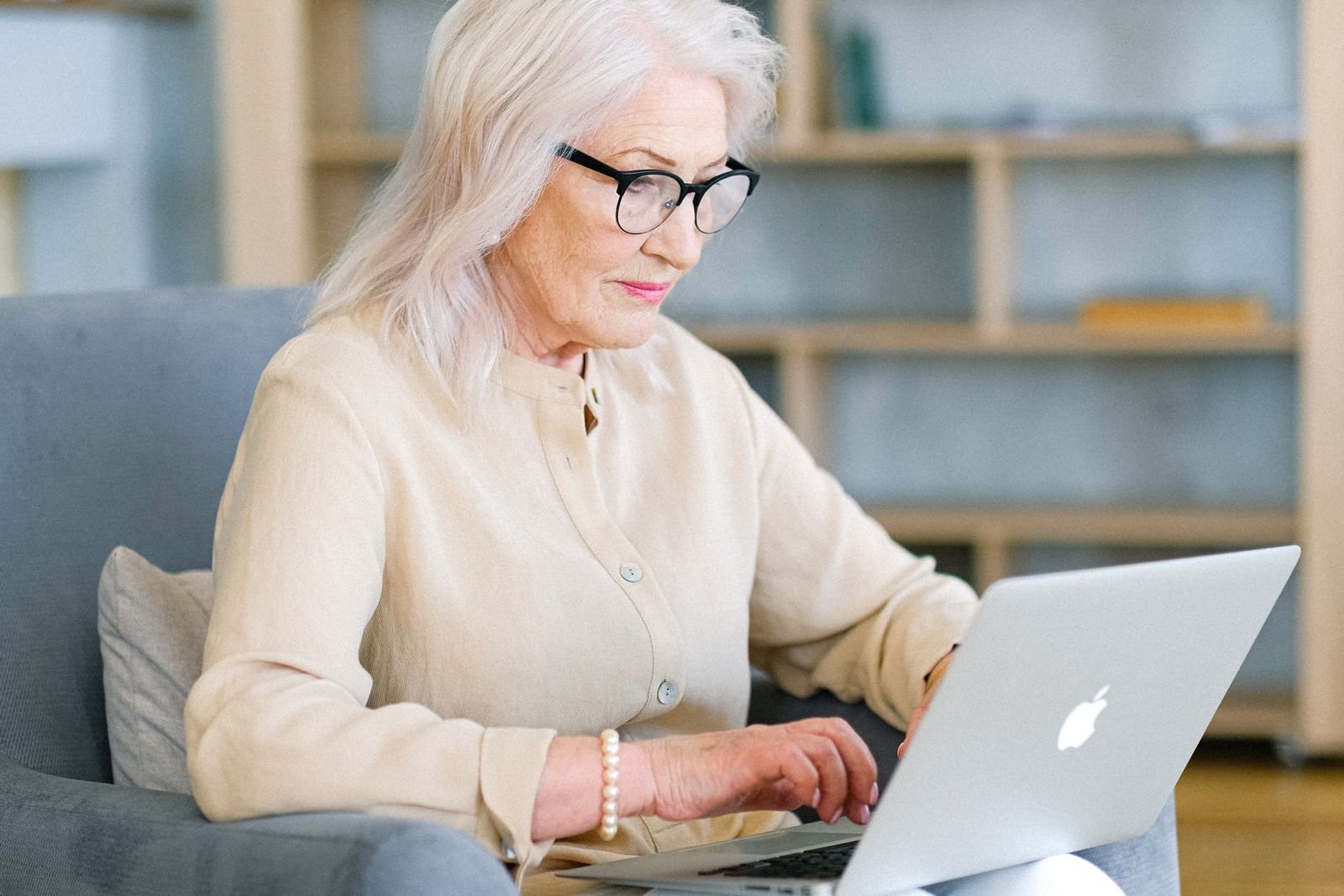 An older woman with white hair and glasses typing on a laptop while seated in an armchair