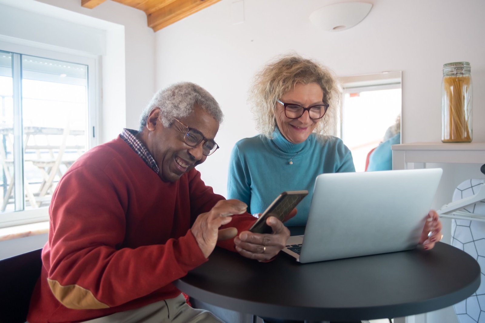 An older man and an older woman sitting at a kitchen table, looking at a phone and a laptop together.